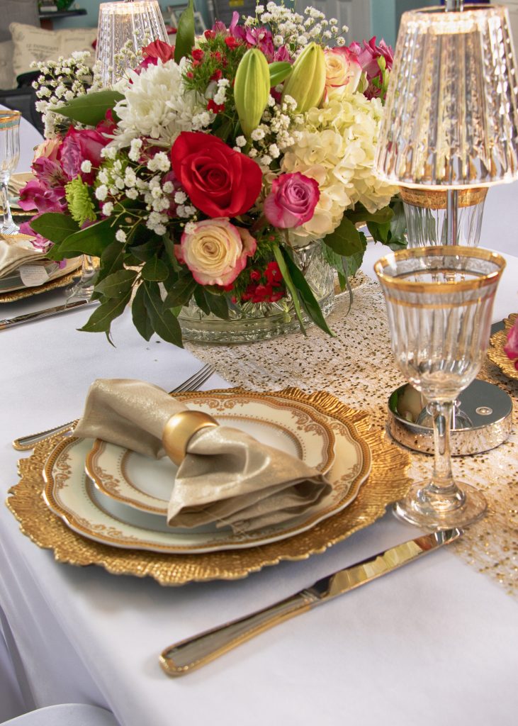 Elegant celebration dinner details featured 24K gold trim dinnerware, glassware, and flatware; linen napkins with rings, in-house floral design in crystal bowl, and silver table lamps.